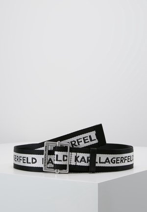 Ceinture en tissu noir et blanc avec texte répétitif "KARL LAGERFELD" et boucle rectangulaire argentée sur une surface blanche.