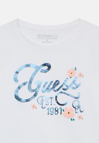 White organic cotton t-shirt featuring shiny blue lettering "Guess EST 1981" with pink and green floral accents.