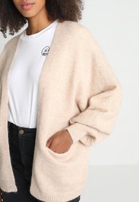 Beige pletený cardigan s oversized rukávy, otevřeným předním dílem a bočními kapsami. Měkká textura s volným střihem, nošený přes bílé tričko.