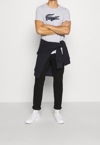 Camiseta gris con gráfico de cocodrilo, pantalones oscuros y zapatillas blancas. Suéter navy atado alrededor de la cintura, ajuste casual, tela suave.