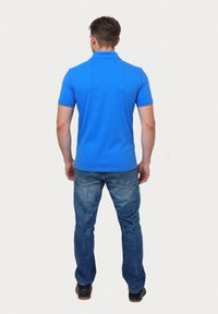 Man som står med ryggen mot kameran, iförd en lysande blå kortärmad polo, blå jeans och mörka skor mot en enkel bakgrund.