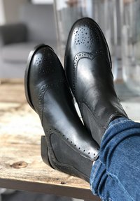 Stivali ankle neri in pelle con dettagli a brogue perforati, texture liscia, pannelli laterali elastici e tacco corto, abbinati a denim.