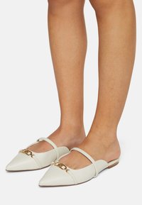 Mules en cuir blanc avec des bouts pointus, présentant un design à double bride et un accent en chaîne dorée à l'avant. Semelle plate. Texture lisse.