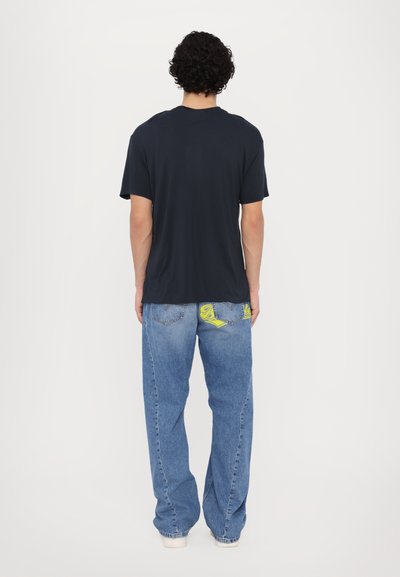 T-shirt bleu marine avec une coupe décontractée, associé à un jean bleu clair présentant des accents jaunes sur les poches arrière. Matériau en coton.