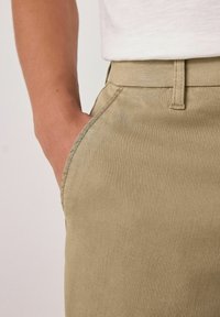 Beige pants with a smooth texture, featuring a side pocket and stitched waistband detail. Close-up view focuses on the fabric and fit.