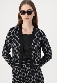 Cardigan noir avec un motif géométrique gris, fermeture à boutons à l'avant et une bordure noire contrastante. Assorti à un haut noir uni.