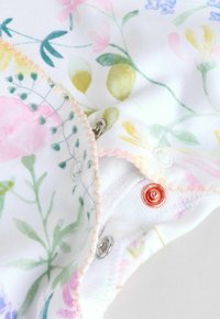 Lähivõte lillemustriga kangast, millel on roosad õmblused ja metallist pressnööbid valgel taustal.