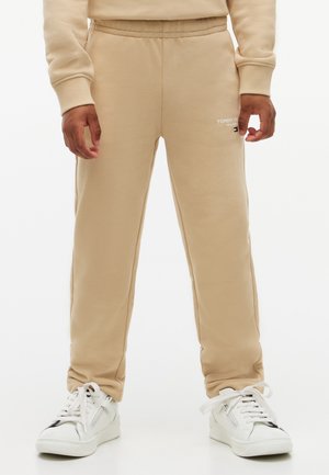 Beige joggingbukser med elastisk talje og hvidt logo på låret, båret med hvide sneakers og matchende beige sweatshirtærmer synlige.