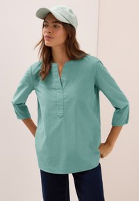 Donna che indossa una blusa verde menta con maniche a tre quarti e un cappellino coordinato, in piedi con le mani sui fianchi su uno sfondo neutro.