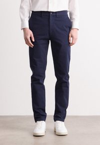 Man som bär smala, marinblå byxor och vita sneakers, som står på ett ljust golv med en enkel bakgrund.