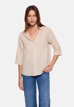 Mujer con camisa beige de botones y mangas tres cuartos, y jeans azules, de pie con una mano en el bolsillo frente a un fondo blanco.