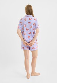 Lilla kortærmet pyjamasæt med røde hjerteprint. Toppen har krave, og shortsene har kontrasterende kant. Blød stoffet.