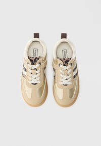 Baskets beiges avec des accents dorés, des lacets blancs et un détail en imprimé léopard. Elles comportent une fermeture éclair et une semelle en caoutchouc texturée pour une meilleure adhérence.