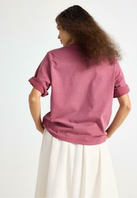 Kortärmad, rosa bomulls-T-shirt med uppvikta ärmar, sedd bakifrån. Kombinerad med en vit kjol, som visar en avslappnad passform och slät textur.