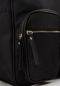 Coin d'un sac en tissu noir avec une petite poche avant zippée équipée d'une fermeture éclair en métal doré et de tirettes en cuir noir.