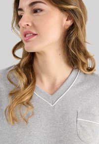 Mujer con cabello ondulado de color castaño claro, vistiendo una blusa gris de cuello en V con ribetes blancos y un bolsillo en el pecho, mirando hacia la izquierda.