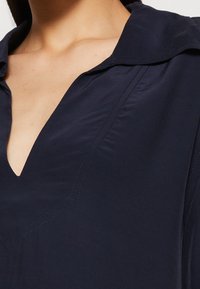 Blouse bleu marine à col, avec un décolleté en V profond, présentant des détails de couture discrets et une texture de tissu lisse. Découpée pour montrer uniquement la partie supérieure.