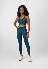 Teal športová podprsenka a legíny s prvkom tie-dye. Materiál je hladký a elastický, ideálny na športové oblečenie. Biele tenisky sú súčasťou.