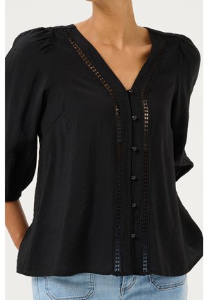 Zwarte V-hals blouse met kanten rand en knopen aan de voorkant, met opgeblazen mouwen en gedragen met lichtblauwe jeans.