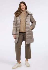 Prošívaný světle hnědý kabát s umělým kožíškem na límci, zipovým zapínáním a bočními kapsami, nošený přes béžový svetr a hnědé kostkované kalhoty s bílými teniskami.