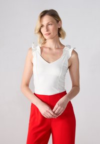 Hvit ermeløs bodysuit med volanger på skuldrene og V-halsdesign, kombinert med høyt liv røde bukser. Jevn tekstur overalt.