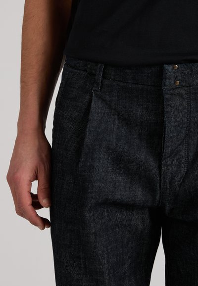 Person wearing dark denim jeans and a black shirt, with their left hand relaxed by their side against a neutral background.