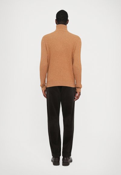 Rjavi rebrasti pulover s turtleneck izrezom in dolgimi rokavi, oblečen z temnimi kordojnimi hlačami in rjavimi čevlji, viden s hrbta.