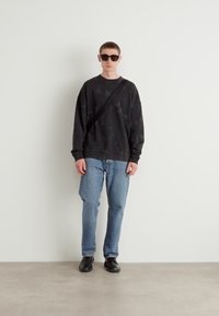 Sudadera negra con patrón, jeans azules, zapatos negros y gafas de sol. El modelo está de pie contra una pared blanca lisa sobre un suelo gris claro.