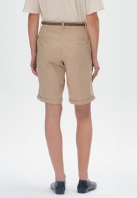 Beżowe szorty z podwiniętymi mankietami, o gładkiej fakturze, z paskiem i dwoma tylnymi kieszeniami, w zestawie z czarnymi butami.