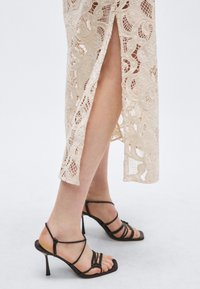 Δαντελένιο φόρεμα σε ανοιχτό μπεζ χρώμα με λεπτομερή σχέδια και πλαϊνή σκίσιμο. Μαύρα πέδιλα με λουράκια, λεπτό τακούνι και διακοσμητικούς κόμπους.