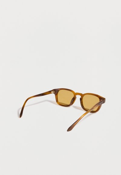 Lunettes de soleil rectangulaires translucides marron avec des verres teintés ambre posées en biais sur une surface blanche.