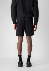Giacca sportiva nera con dettagli a strisce, abbinata a pantaloni corti neri. Modello che indossa calze e scarpe nere, visto da dietro.
