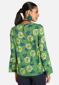 Eine Frau mit langen, zurückgebundenen Haaren trägt eine grüne Bluse mit floralen Mustern, die einen stilvollen Rücken-Ausschnitt zeigt, ergänzt durch blaue Accessoires.