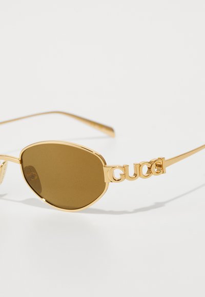 Kultaiset kehykset aurinkolaseille, joissa on ruskeat linssit ja "GUCCI"-logo sisällytettynä varren muotoiluun valkoisella taustalla.