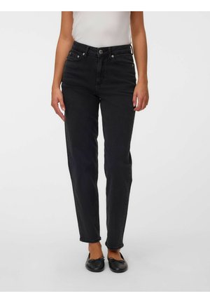 Femme portant un jean noir taille haute, un top blanc et des ballerines noires, debout devant un fond gris uni.