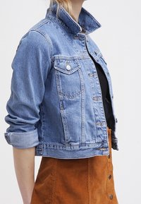 Kvinna som bär en ljusblå denimjacka med uppvikt ärmar över en svart topp och en brun knappad kjol, stående mot en vit bakgrund.