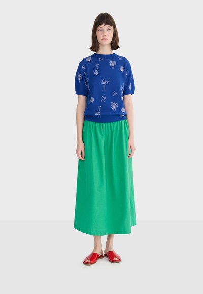 Kobieta stojąca, ubrana w niebieską bluzkę z krótkim rękawem, ozdobioną białymi kwiatowymi i ptasimi wzorami, długą zieloną spódnicę oraz czerwone, otwarte sandały na płaskim obcasie.