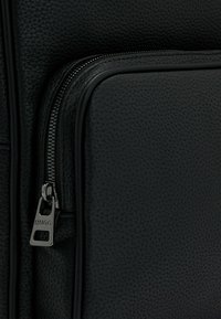 Black textured leather bag with a prominent zipper. Features a silver hardware pull tab labeled "HUGO." Smooth, matte accents and structured design.