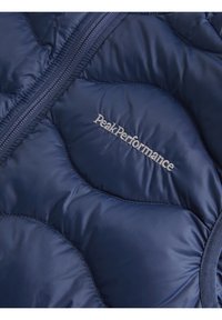 Tamno plava puffer jakna s kviltanim dizajnom, glatkom teksturom i srebrnim logotipom "PeakPerformance" na prsima. Vidljiv detalj sa zatvaračem.