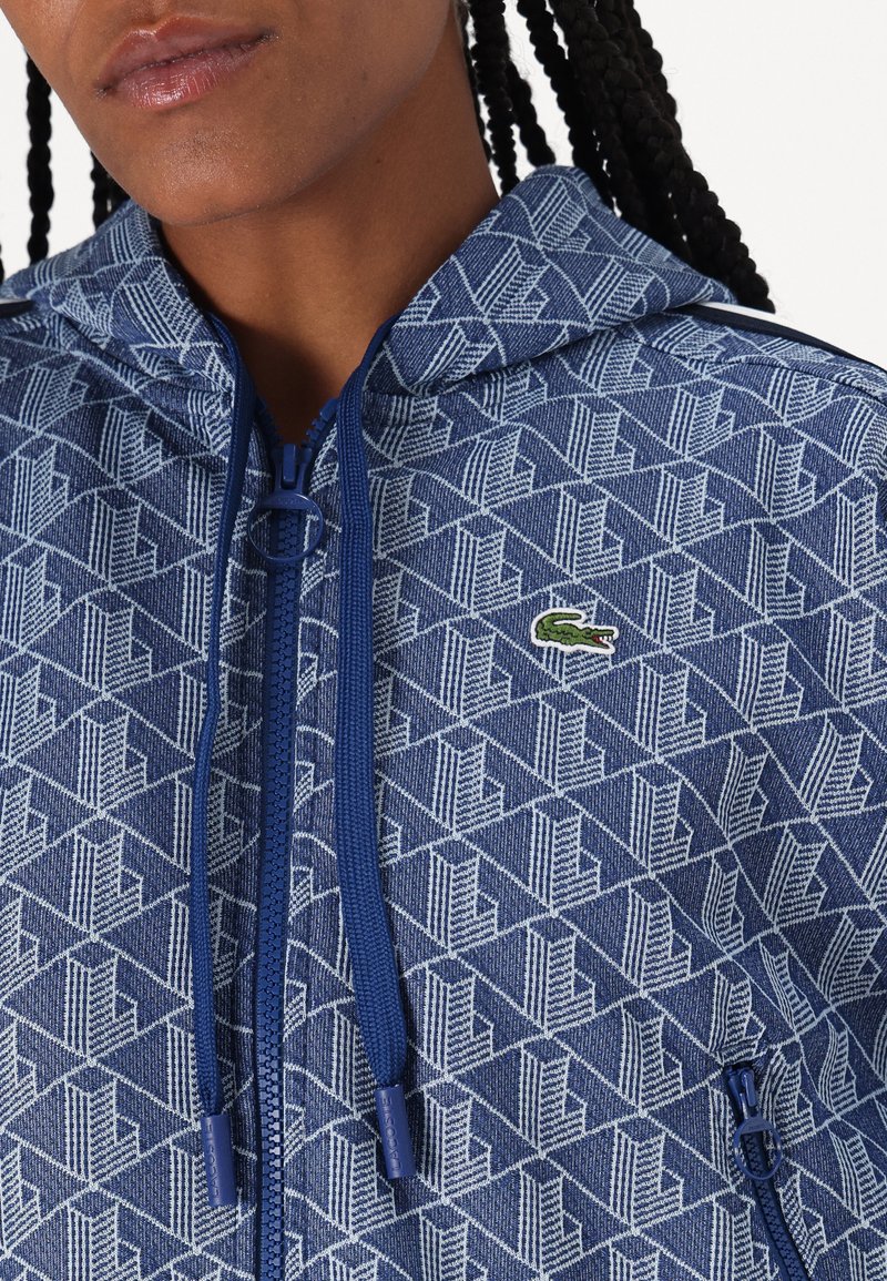 Felpa blu con motivo geometrico. Presenta una zip frontale, cordini e un piccolo logo verde come accento. La texture sembra a maglia.