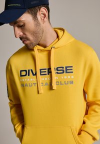 Żółta bawełniana bluza z kapturem z kieszenią kangurką, ma napis "DIVERSE NAUTICAL CLUB" w granatowym i białym kolorze. Sznureczki są fakturowane.