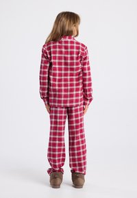 Kind staat met de rug naar de camera, in rode en witte gestreepte pyjama's en bruine fluffy slippers, tegen een effen witte achtergrond.