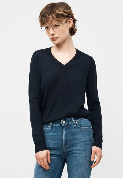 Femme aux cheveux bruns courts portant des lunettes en métal fines, un pull noir à col en V et un jean bleu, sur un fond clair uni.