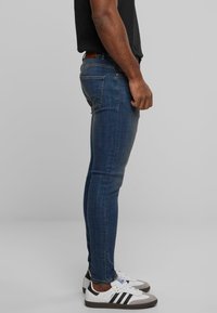 Jeans de mezclilla azul ajustados con una textura lisa, cinco bolsillos y un sutil patrón desvanecido. Combinados con zapatillas deportivas blancas con rayas negras.