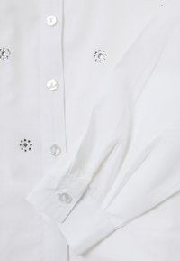 Camicia bianca con bottoni perlacei e dettagli in strass decorativi sul davanti. Tessuto morbido con maniche lunghe.