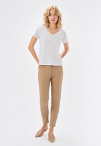 Młoda kobieta stoi z rękami za plecami, ubrana w białą koszulkę z dekoltem w serek, beżowe wąskie spodnie i beżowe buty na obcasie.