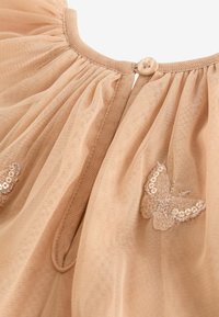 Robe en tulle beige avec un décolleté rond, ornée de plis et de deux motifs de papillons brodés avec des sequins. Fermeture au dos par des boutons.