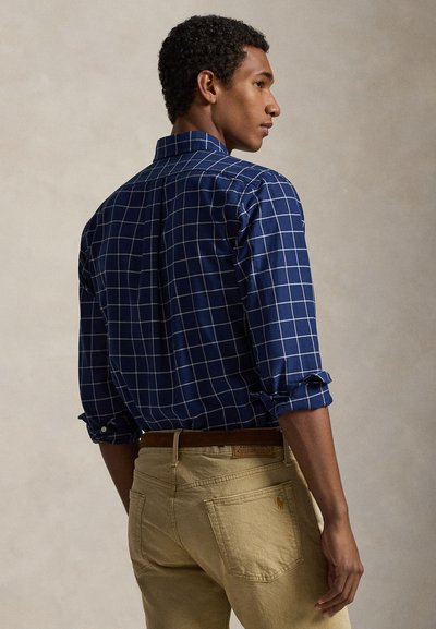 Navy blue checkered shirt with rolled sleeves, made of lightweight fabric, worn with tan pants, featuring a brown belt with logo detailing.