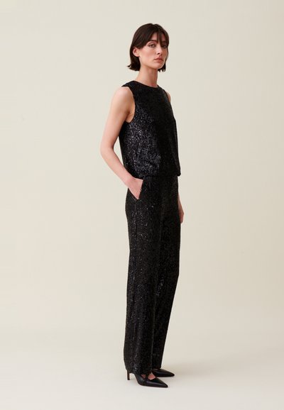 Mono negro con lentejuelas, de tirantes y diseño de pantalón de pierna ancha. Textura suave con acabado brillante. Incluye bolsillos laterales y tacones puntiagudos.