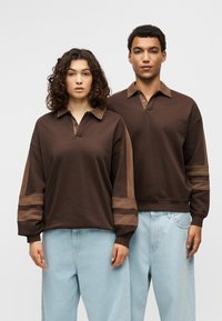 Καφέ πλεκτά πόλο με αντιθετικά μανίκια, γιακά με κουμπιά και άνετη εφαρμογή. Φοριέται με ανοιχτό μπλε φαρδιά τζιν.